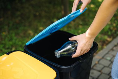 Yakın plan kadın portresi geri dönüşüm kutusuna boş cam şişe atıyor. Parktaki plastik kutuların farklı renkleri.