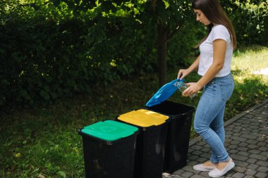 Geri dönüşüm kutusuna boş cam şişe atan kadın portresi. Parktaki plastik kutuların farklı renkleri.
