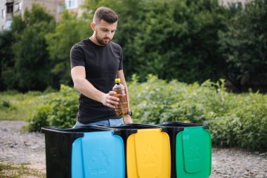 İnsan elinin geri dönüşüm kutusuna boş plastik su şişesini attığı yakın plan. Ağaç geri dönüşüm kutuları açık havada.