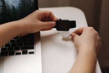 Kadın eli, kablosuz şarj cihazına akıllı saat taktı. Laptopun yanındaki iş yerinde..