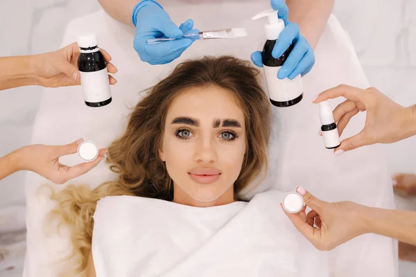 Top view of beautiful woman with cosmetology tools lying on the medical couch. Facial treatment concept. Hands of three beautucian hold different tools.