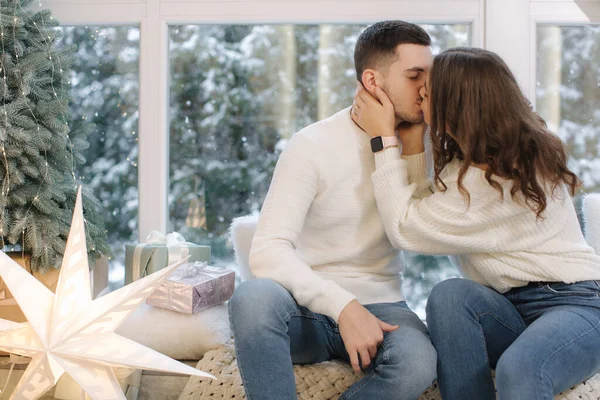 Beautiful young couple sitting in front of big window and fir tree. Its snow outside.