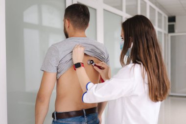 Stetoskop kullanarak klinikteki bir hastanın akciğerlerini kontrol eden tıbbi maskeli kadın bir doktor. Tıbbi muayene. Arka plan.