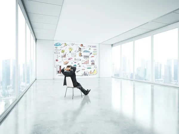 Businessman sitting in empty bright office with business strategy on the wall — Stock Photo, Image