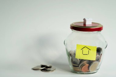 Ev alabilmek için para biriktirme kavramı. İkisi de cam kavanozda, kapağında ev ve kavanozun dışında madeni paralar var. Fotoğraf: 17 Ocak 2022 Stockholm, İsveç 