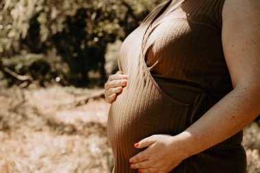 Yakında bebek sahibi olma umuduyla neşeli bir hamilelik. Tanımlanamayan hamile bir kadının açık havada, göbeğinde elleri olan sıradan bir elbiseyle çekilmiş yakın plan fotoğrafı..