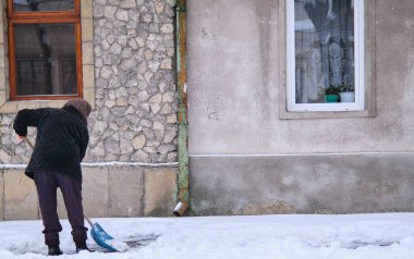 Sıcak ceketli yaşlı kadın kar yığınlarını kaldırımda kürekle temizliyor.