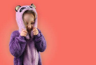 Küçük kız çocuğu pembe arka planda gülümseyen kapüşonlu mor pijama giyiyor.