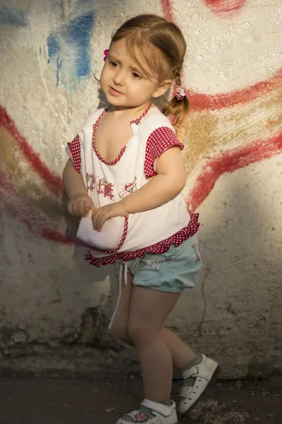 Plachý dítě. Dívka ve věku 2-3 let s copánky na vlasy nebo s ponocem stojící nedaleko barevné kamenné zdi — Stock fotografie