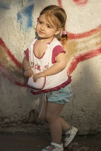 Shy child. Girl aged 2-3 years with hair braids or ponytails standing near a colored stone wall — Stock Photo, Image