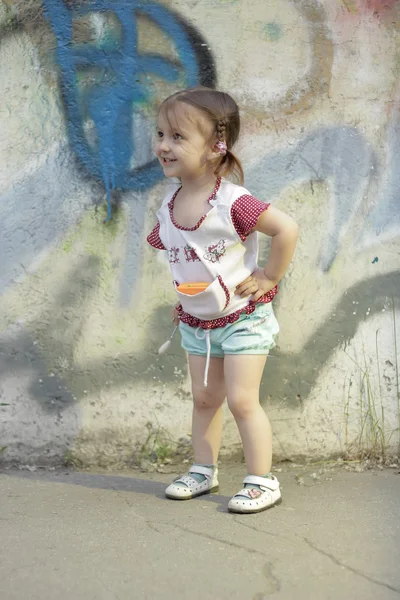 Positive kid. Happy little girl 2-3-4 years old with braids on her head, stands and smiles in the street near a concrete wall with graffiti, the girl's hands on her hips. — Stock Photo, Image