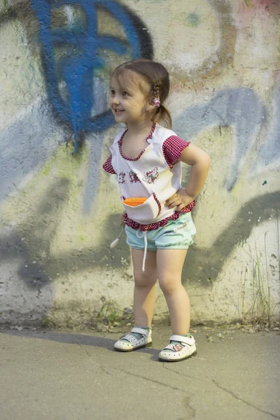 Positive kids. Happy little girl 2-3-4 years old with braids on her head, stands and smiles in the street near a concrete wall with graffiti, the girl's hands on her hips. — Stock Photo, Image