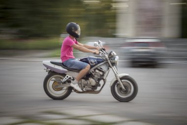 Motosiklete binen adam. Pembe tişörtlü ve kot şortlu genç adam, kafasında motosiklet kaskı olan genç adam sokakta motosiklete hızla biniyor.