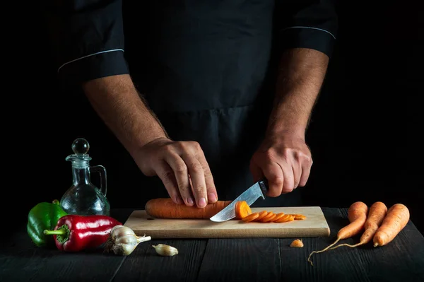 Professional chef is cutting carrots for vegetable soup in the restaurant kitchen. Close-up of the hands of cook during work. Carrot die