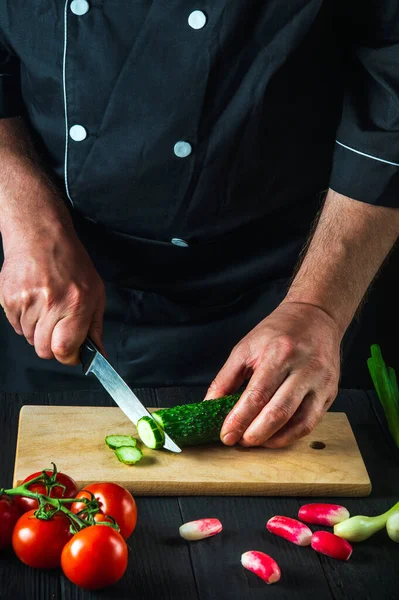 Professional chef cuts a green cucumber on a restaurant kitchen cutting board for salad. Vegetable diet or snack idea.