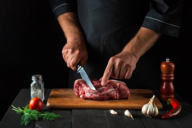 Aşçı mutfakta bıçakla et keser, yemek hazırlar. Bir restoranda lezzetli bir öğle yemeği hazırlamak için mutfak masasındaki sebze ve baharatlar..