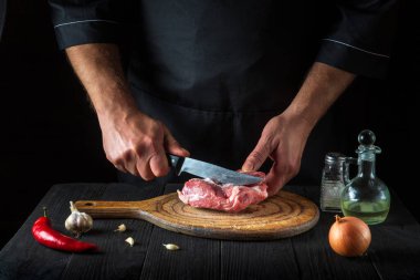 Aşçı mutfakta bıçakla et keser, yemek hazırlar. Bir restoranda lezzetli bir öğle yemeği hazırlamak için mutfak masasında sebzeler ve baharatlar..