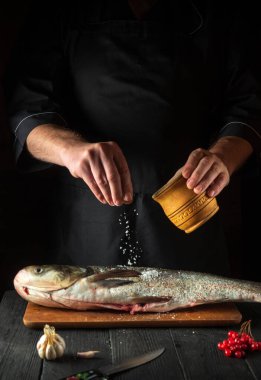 Aşçı restoranın mutfağında gümüş sazan balığı hazırlıyor. Aşçı balığın üzerine tuz serper. Mutfak masasında çalışma ortamı