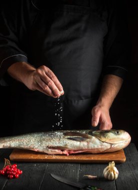 Aşçı mutfakta gümüş sazan balığı hazırlıyor. Şef balığın üzerine tuz serper. Mutfak masasında çalışma ortamı.