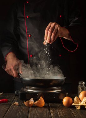 Profesyonel şef tavada yumurta pişirirken tuz ekler. Mutfak masasında çalışma ortamı. Restoran ya da otel için menü fikri.