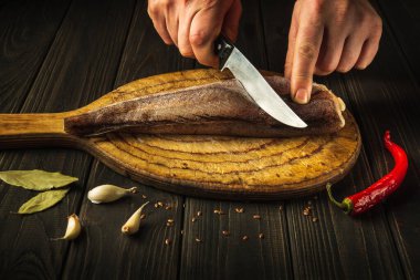 Aşçı, tahta bir kesme tahtasıyla taze Merluccius balığını bıçakla keser. Eski bir tarife göre lezzetli bir balık yemeğinin ev yemeği..