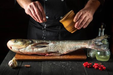Aşçı restoranın mutfağında gümüş sazan balığı hazırlıyor. Aşçı balığın üzerine tuz serper. Mutfak masasında çalışma ortamı