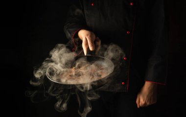 Şef, siyah arka planda buharlı bir tavada yemek hazırlıyor. Restoran ve otel servisi kavramı. Asya mutfağı.