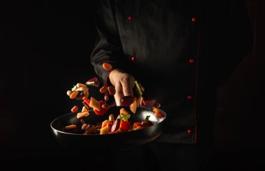 Profesyonel şef taze sebzeleri kızartma tavasında hazırlar. Büyük mutfak ve yemek tarifi koyu arka planda. Ücretsiz reklam alanı.