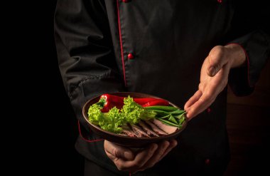 Profesyonel şefin elinde bir tabak doğranmış biftek ve sebze var. Karanlık arka planda yemek pişirme kavramı. Asya mutfağı
