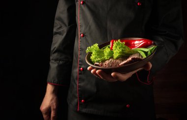 Şefin elinde et ve sebzelerle dolu bir tabak var. Karanlık arka planda yemek hazırlama konsepti. Asya mutfağı