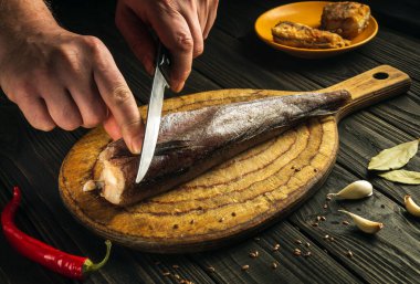 Aşçı, tahta kesme tahtasıyla taze Merluccius balığını bıçakla keser. Eski bir tarife göre lezzetli bir balık yemeğinin ev yemeği.