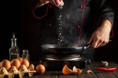 Şef tavada yumurta pişirirken tuz ekler. Mutfak masasında çalışma ortamı. Restoran ya da otel için menü fikri
