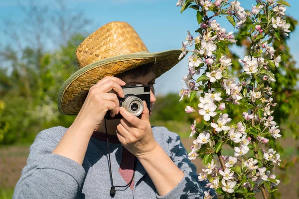 庭のリンゴの木の花の枝を撮影する古いカメラと藁帽子の中年女性の肖像画 — ストック写真