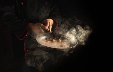 Şef siyah arka planda buharlı bir tavada yemek hazırlıyor. Restoran ve otel servisi kavramı