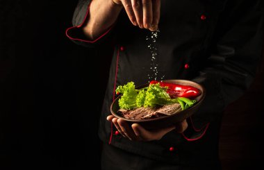Şef dilimlenmiş bifteğe tuz serpiyor ve bir tabakta biftek ve sebzeler var. Yemek servisi konsepti..