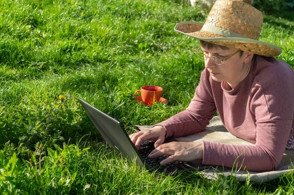 A forty-five-year-old woman in glasses works as a freelancer outdoors. Rest with a laptop in nature in the park on a sunny day against the backdrop of green grass.
