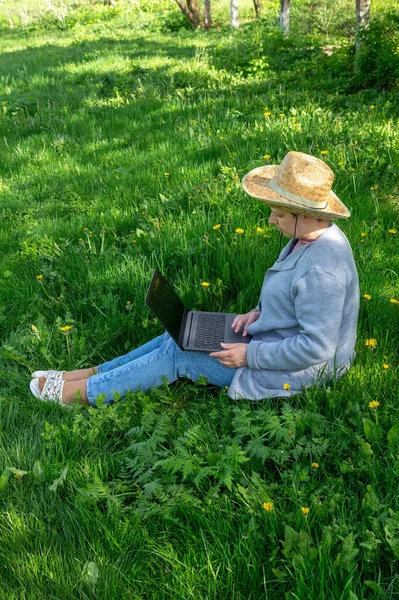 Rest with a laptop in nature in the park on a sunny day against the backdrop of green grass. Woman in her forties freelancer outdoors.