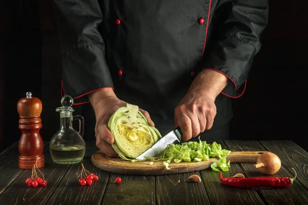 Professional chef cuts fresh cabbage with knife for salad on a kitchen table with fresh vegetables. Cooking and restaurant or hotel concept