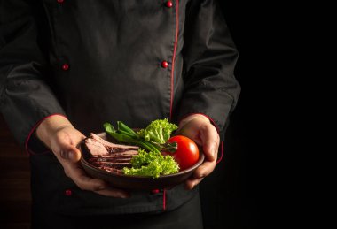 Şef bir tabak dilimlenmiş biftek ve sebze servis ediyor. Karanlık arka planda yemek pişirme kavramı..