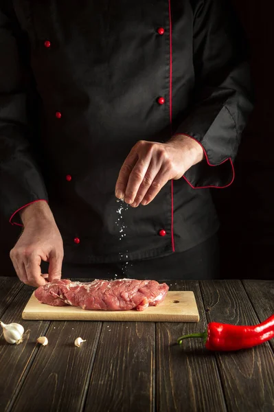 Professional chef sprinkles raw meat with salt. Preparing beef meat before baking. Working environment in the kitchen