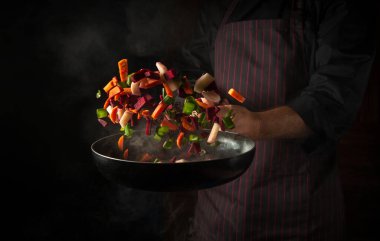 The hand of a professional chef throws pieces of vegetables into a hot frying pan with steam on a black background. The concept of cooking in the hotel. Free ad space