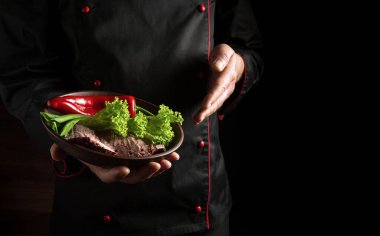 Şef, dilimlenmiş et ve sebzeli bir tabak saklar. Karanlık arka planda yemek pişirme kavramı.
