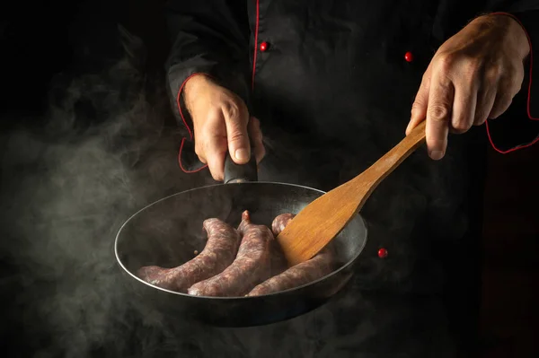 The chef is frying sausages for hot dogs in a frying pan. Close-up of a cook hands with a hot pan in the kitchen. Free space for advertising