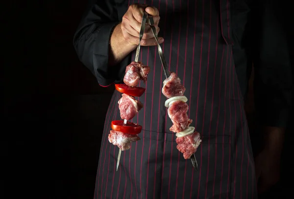 The chef holds shish kebabs with raw lamb meat on skewers in his hands. Asian national cuisine