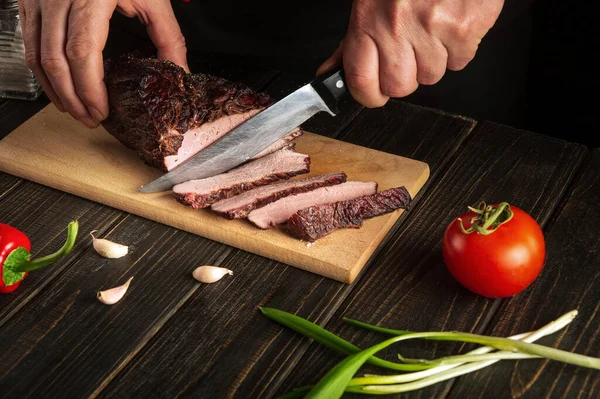 Master chef cuts the baked beef meat on a cutting board. The idea of preparing a delicious lunch