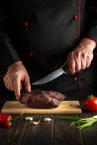 Professional chef cuts the baked beef steak with a knife on a wooden board. The concept of the cooking process food for hotel