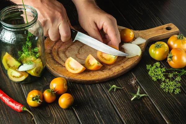 Cooking pickled vegetables in a jar. The cook cuts tomatoes with a knife on a cutting board in the kitchen. Close-up of chef hands while working.