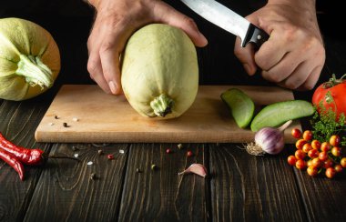 Slicing vegetable marrow with a knife before cooking by the hands of the cook on a wooden cutting board. In cooking, the thick flesh of zucchini is used.