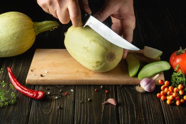 Hands of a cook with a knife cutting vegetable marrow on a wooden cutting board before preparing vegetarian food