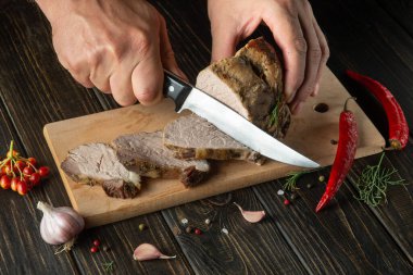The cook cuts the grilled veal meat into small slices. Work environment on the kitchen table with condiments or spices. Black or dark background with space for recipe or menu